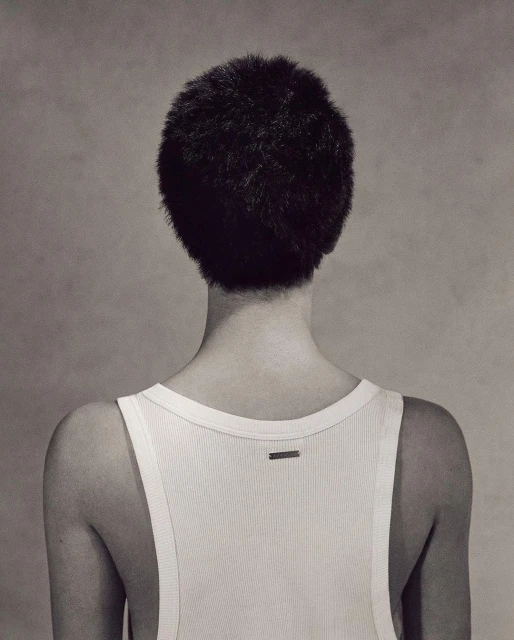 Back view of a person with short, dark hair wearing a white ribbed tank top against a neutral grey backdrop in a studio shot.