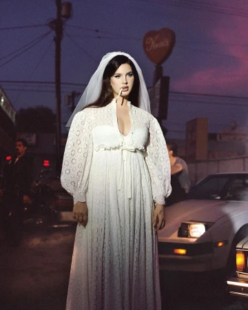 Woman in a wedding dress stands smoking a cigarette against a cityscape background with cars and a building with a heart-shaped sign.