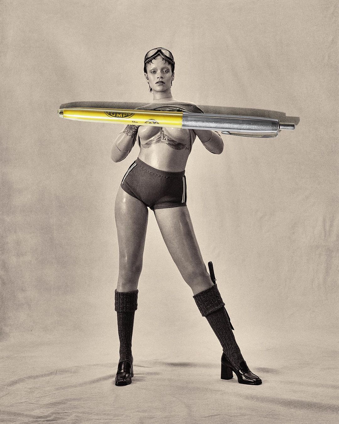 A model poses with a very large yellow marker in a studio setting, wearing sports bra and shorts, goggles and chunky knit boots.