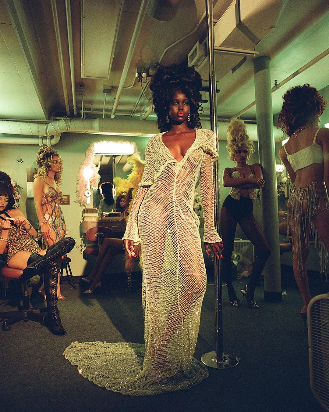 Glamorous image showcases an African-American woman in a sheer gown, backed by a group of women in a dressing room with vintage decor and lighting.