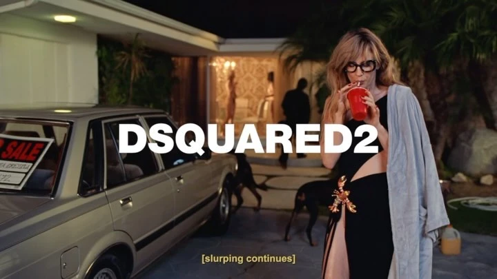 A woman in a black dress sips from a red cup against the backdrop of a house, a car, and other people.