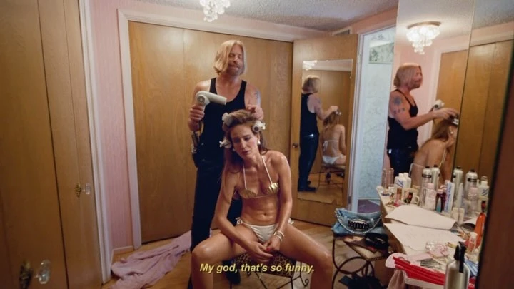 Indoor scene of a man using a hair dryer on a woman in a gold bikini, seated on a stool in front of a vanity in a bedroom.