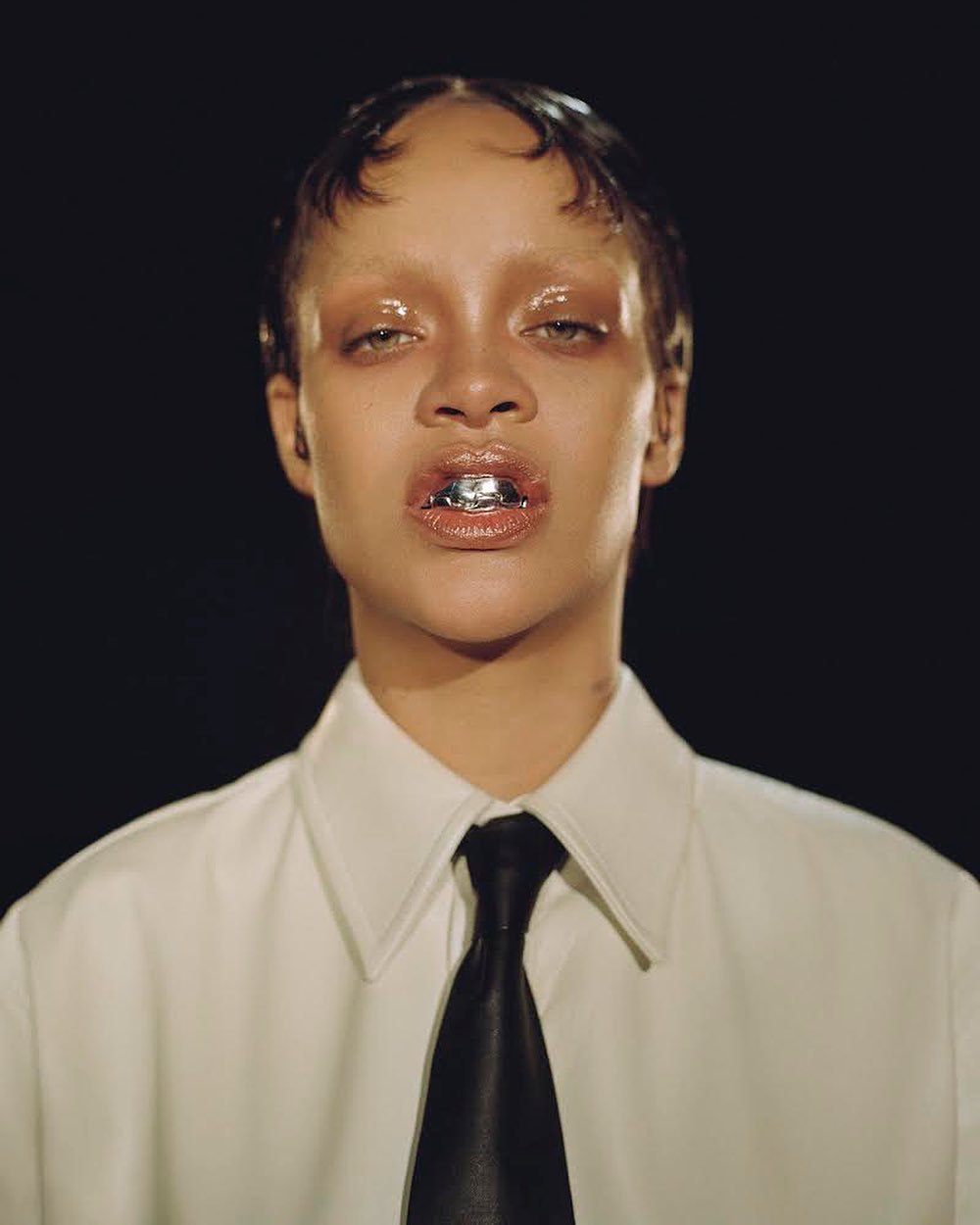 Close-up portrait of a woman with tan skin, wearing a white shirt and black tie, with a silver grill on her teeth, against a dark background.