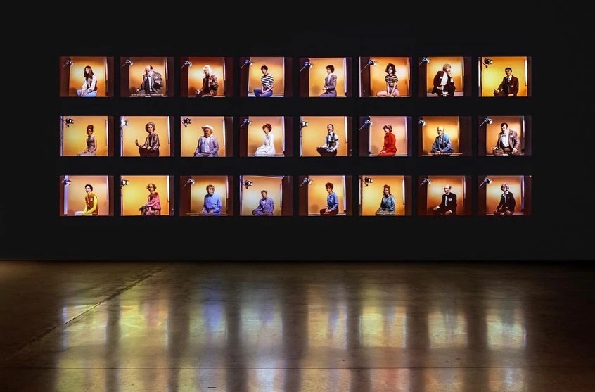 A grid of monitors shows people in seated meditation poses against warm backgrounds in a dark gallery, creating a contemplative atmosphere.