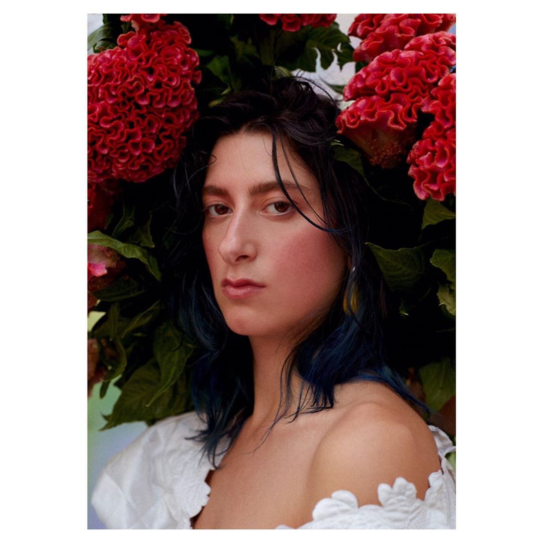 Portrait of a woman with dark hair, wearing a white top, posing in front of red flowers with green leaves, eye-level angle.