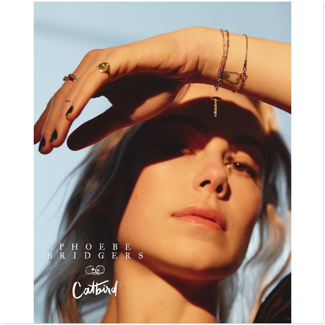 Close-up portrait of Phoebe Bridgers wearing gold jewellery and holding her hand across her forehead under warm sunlight.