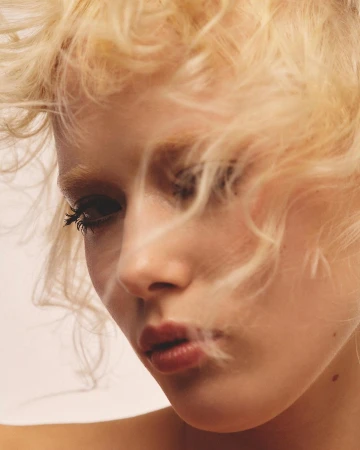 Close-up portrait of a young woman with curly blonde hair blowing across her face and dark mascara.