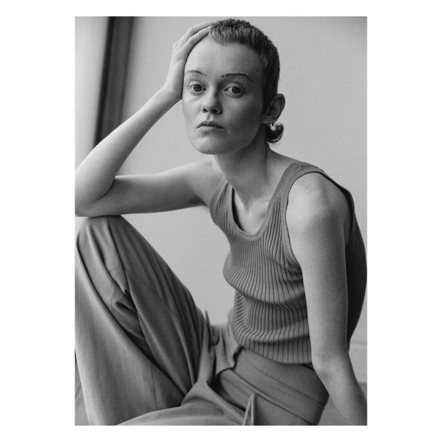 Monochrome portrait of a young adult female with a buzzcut, seated with one arm raised to her head, wearing a tank top and trousers.