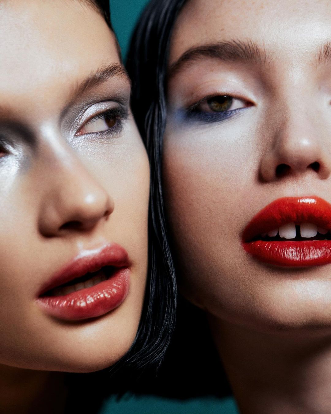 Close-up beauty shot of two women with bold makeup featuring red lipstick and silver and blue eyeshadow on a teal backdrop.