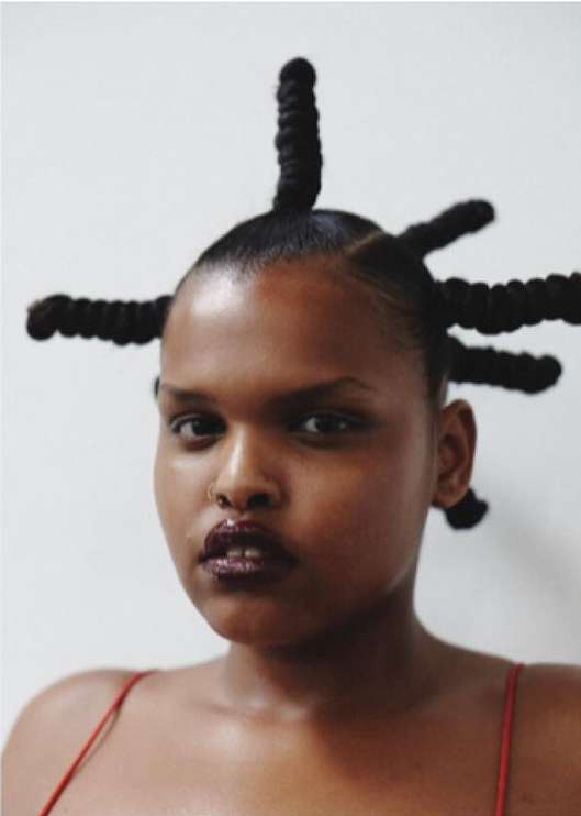 Close-up portrait of a Black woman with sculptural twisted updos and dark glossy lipstick against a neutral wall