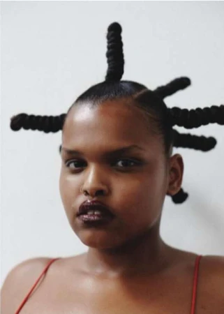 Close-up portrait of a Black woman with sculptural twisted updos and dark glossy lipstick against a neutral wall