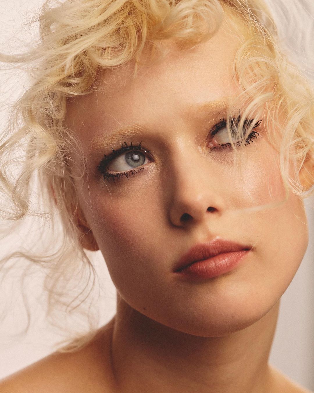 Close-up portrait of a blonde female model with curly hair looking up and away, light makeup, shot indoors against a bright neutral backdrop.