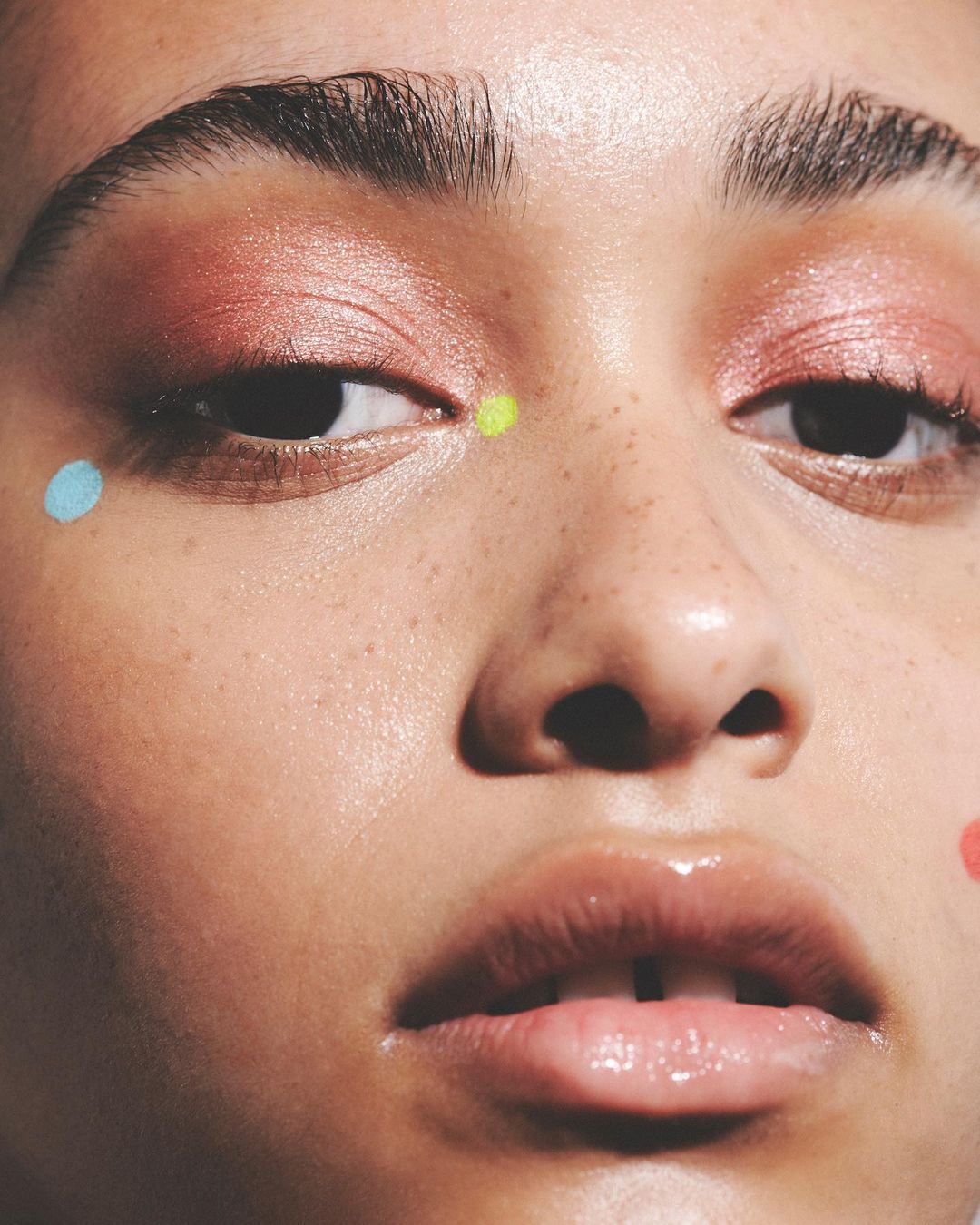 Close-up of a young adult female's face with coral eyeshadow and blue, green, and coral dots on the skin. Beauty shot.