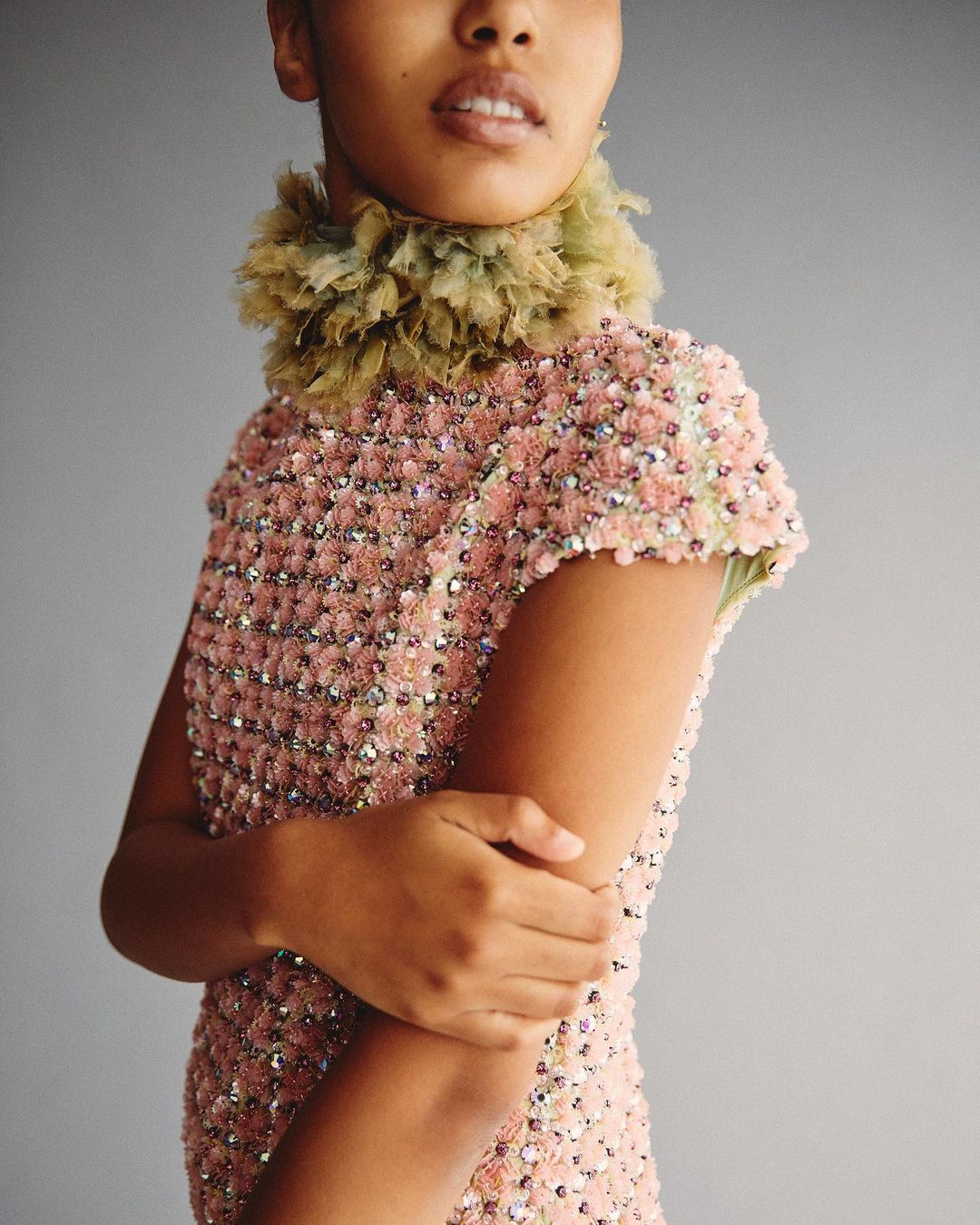 Portrait of a young woman with olive skin wearing a light pink beaded dress and a feathered collar against a gray background.