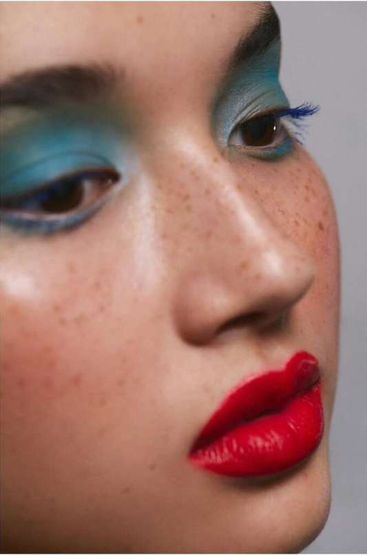 Close-up portrait of a woman with bright makeup, featuring red lipstick, turquoise eyeshadow, and freckles, against a neutral background.