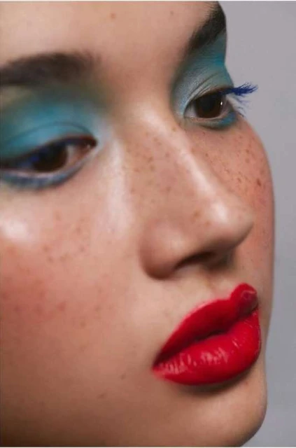 Close-up portrait of a woman with bright makeup, featuring red lipstick, turquoise eyeshadow, and freckles, against a neutral background.