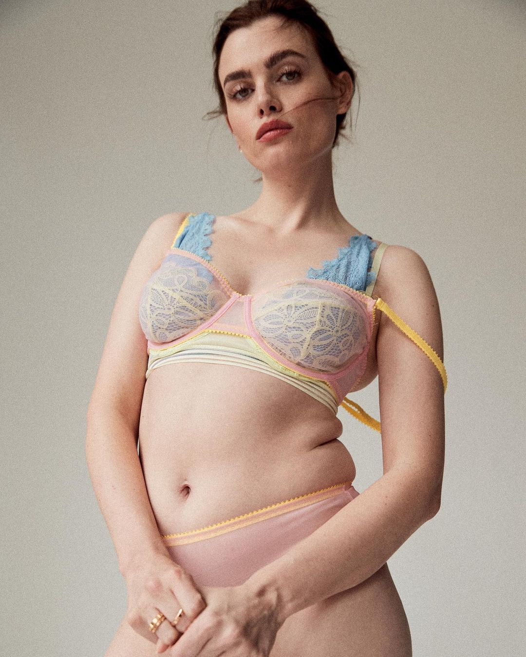 Woman poses in pastel blue, yellow, and pink lingerie set against a plain light gray background in a medium studio shot.