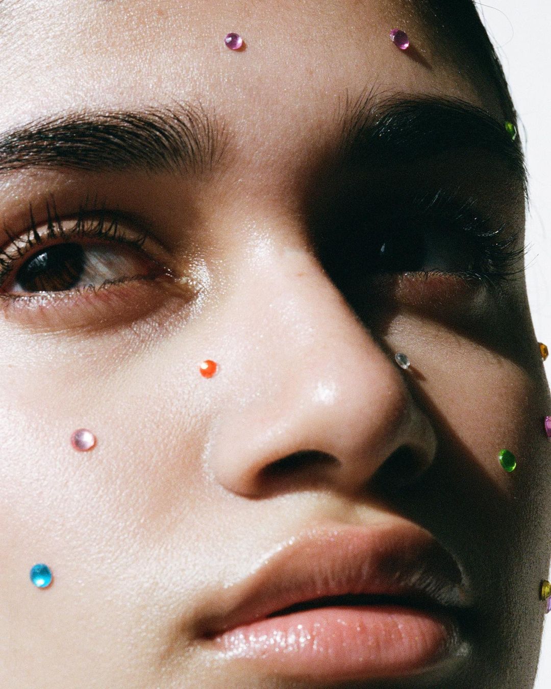 Close-up portrait of a woman's face with colorful rhinestones, emphasizing her smooth skin and the artistic makeup detail against a light background.