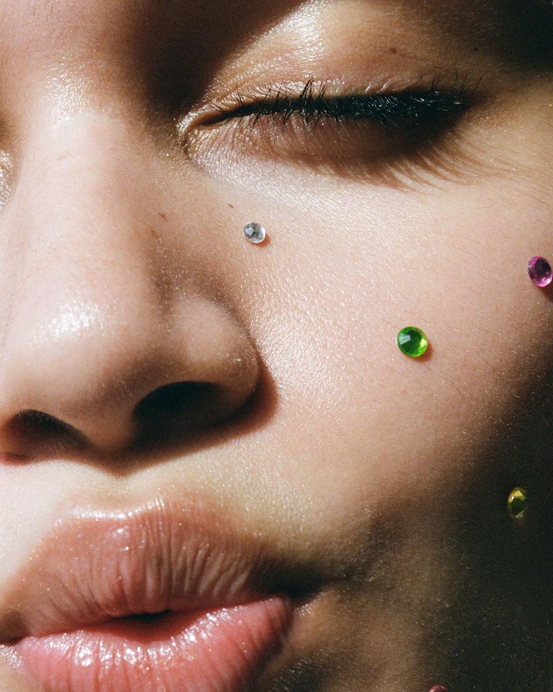 Close-up of a young adult female face with light skin, decorated with small green, pink, and silver gems, partially shaded by light.