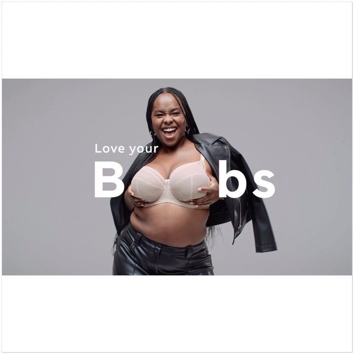 Smiling adult female in a bra and leather jacket poses in front of a grey background with the words 'Love your B**bs' on screen.