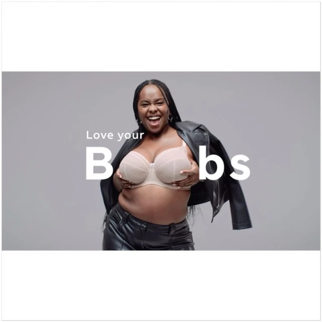 Smiling adult female in a bra and leather jacket poses in front of a grey background with the words 'Love your B**bs' on screen.