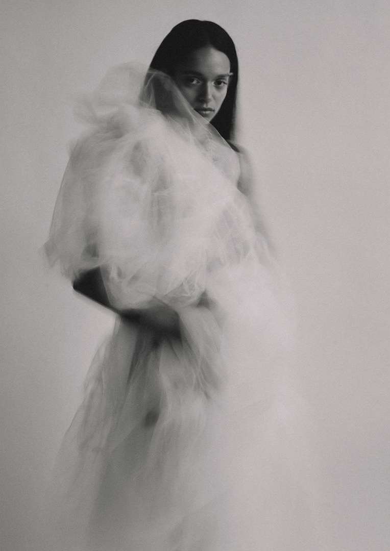 Monochrome portrait of a woman in a white tulle dress, looking at the camera with a serious expression against a light grey background.