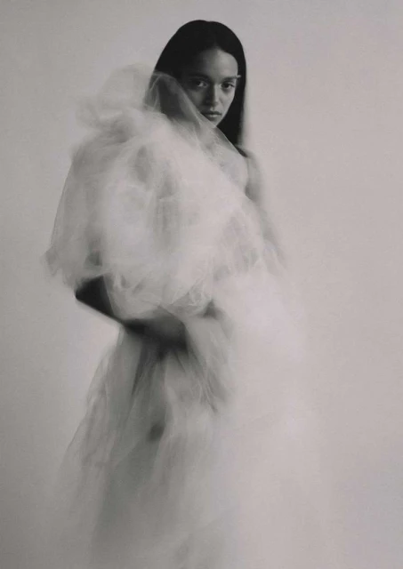 Monochrome portrait of a woman in a white tulle dress, looking at the camera with a serious expression against a light grey background.