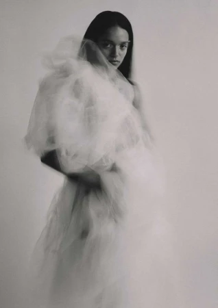 Monochrome portrait of a woman in a white tulle dress, looking at the camera with a serious expression against a light grey background.