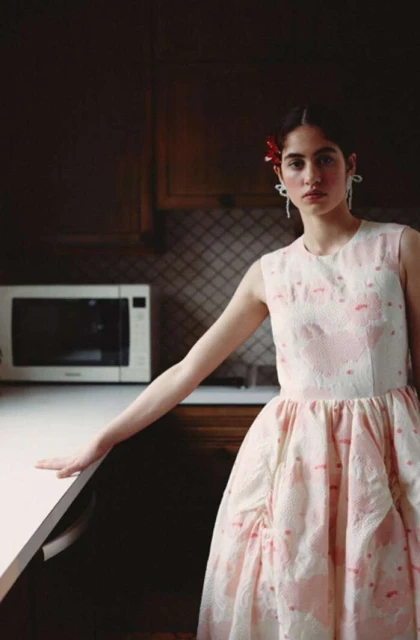 Woman poses in pink and white dress inside kitchen with dark cabinets, white countertops, and a microwave in the background.