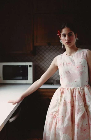 Woman poses in pink and white dress inside kitchen with dark cabinets, white countertops, and a microwave in the background.