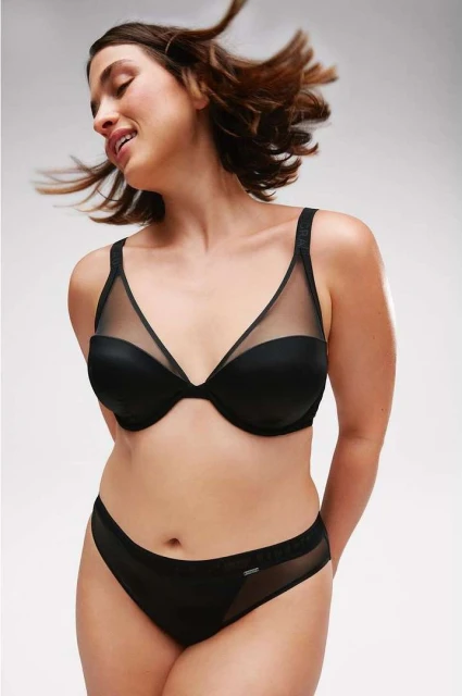 A woman poses in black lingerie against a soft white background, showcasing the bra and panty set in a simple, elegant portrait.