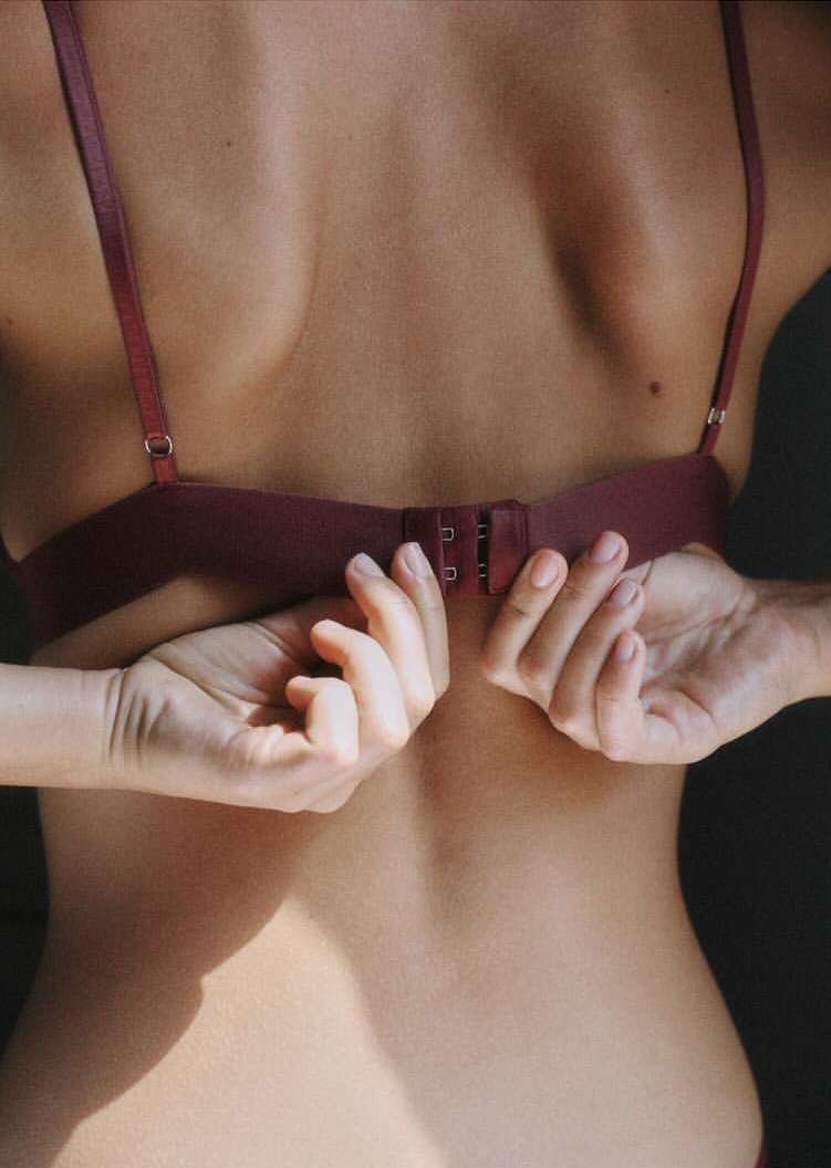 Close-up shot of an adult female's back as she fastens her bra, capturing the intimate moment in soft lighting indoors.