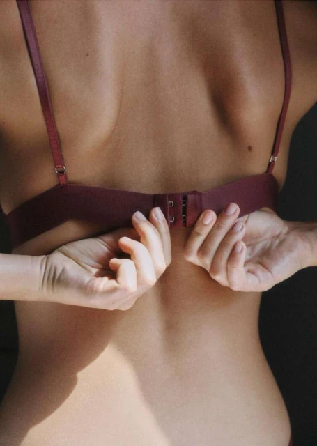Close-up shot of an adult female's back as she fastens her bra, capturing the intimate moment in soft lighting indoors.