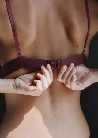 Close-up shot of an adult female's back as she fastens her bra, capturing the intimate moment in soft lighting indoors.