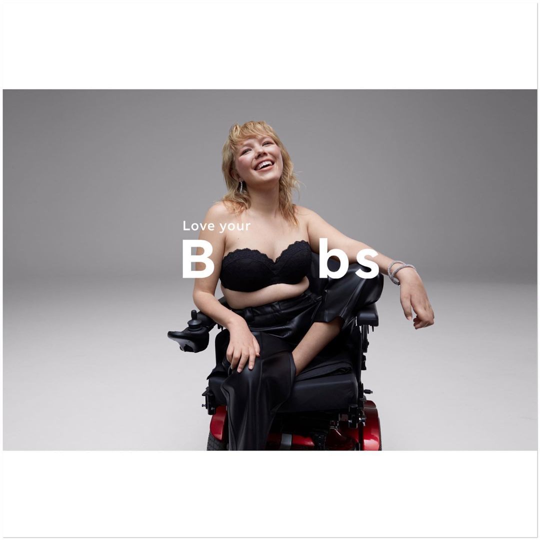 Smiling woman in black lingerie and pants sits in a wheelchair in a studio. Gray background. The text 'Love your B bs' is visible.