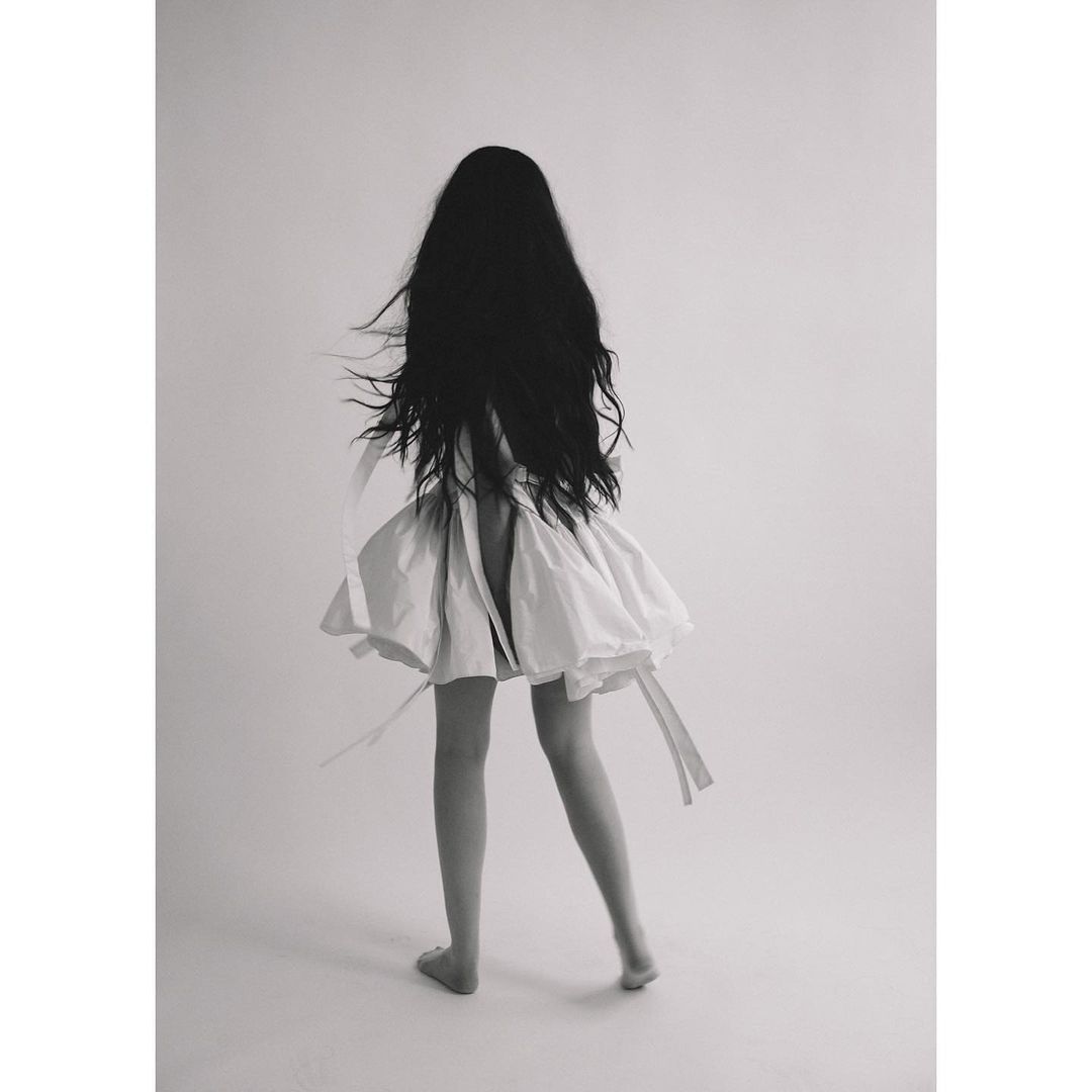 Monochrome portrait from the back of a slender woman with long black hair in a white dress against a clean studio backdrop.