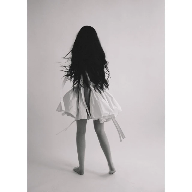 Monochrome portrait from the back of a slender woman with long black hair in a white dress against a clean studio backdrop.