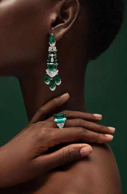 Close-up of a dark-skinned adult female showcasing statement emerald earrings and a cocktail ring against a dark green backdrop.