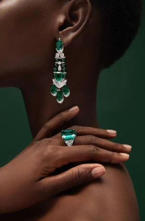 Close-up of a dark-skinned adult female showcasing statement emerald earrings and a cocktail ring against a dark green backdrop.