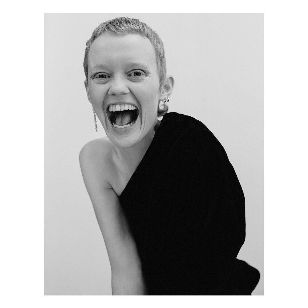 Black and white portrait of a fair-skinned woman with a buzzcut smiling widely, wearing a black garment against a neutral background.