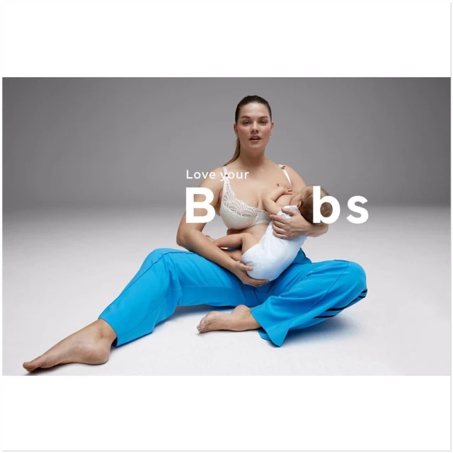 A woman wearing a bra and blue pants sits cross-legged on the floor and breastfeeds a baby on a grey studio background.