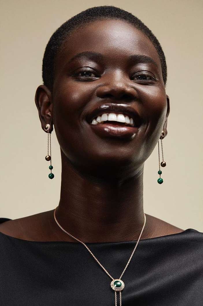 Elegant portrait of a Black woman smiling with dangling earrings and a pendant necklace in a studio setting with a cream background.