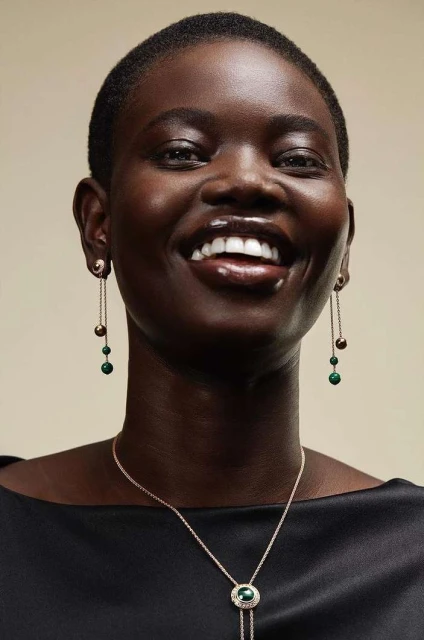 Elegant portrait of a Black woman smiling with dangling earrings and a pendant necklace in a studio setting with a cream background.