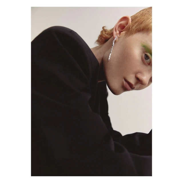 Close-up fashion shot of a female model with blonde hair, wearing an elegant black jacket and long dangling earrings on a white background.
