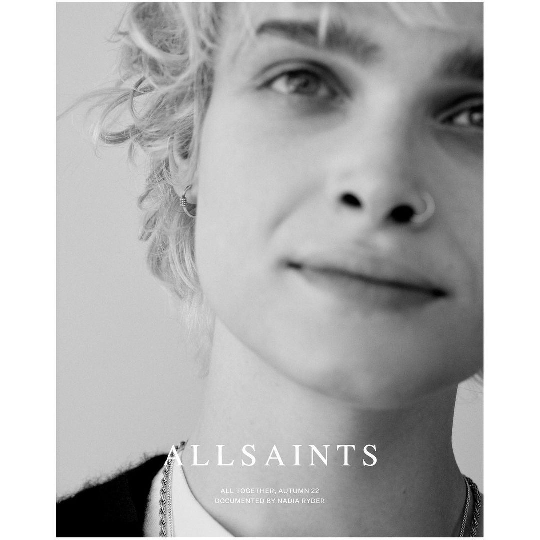 Close-up black and white portrait of a young adult with blonde hair, wearing jewelry and a black shirt against a light background.