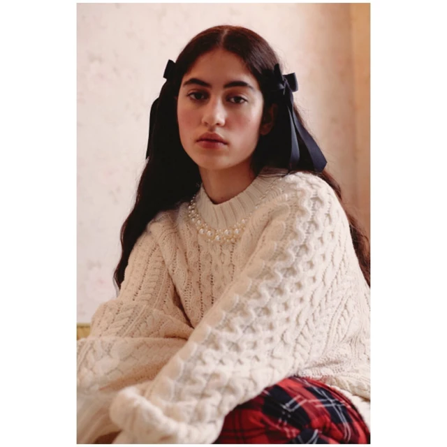Portrait of a young woman with long brown hair and navy hair ribbons, wearing a cream-colored cable knit sweater and pearl necklace indoors.
