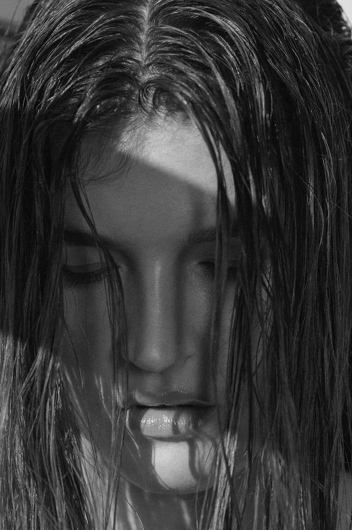 Monochrome portrait of a young woman with wet hair partially covering her face, highlighting her features in soft, diffused light.
