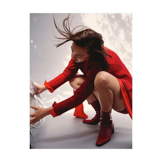 Woman in a red blazer and matching boots is crouched down near a vintage fan indoors with a white wall background in a fashion style photograph.