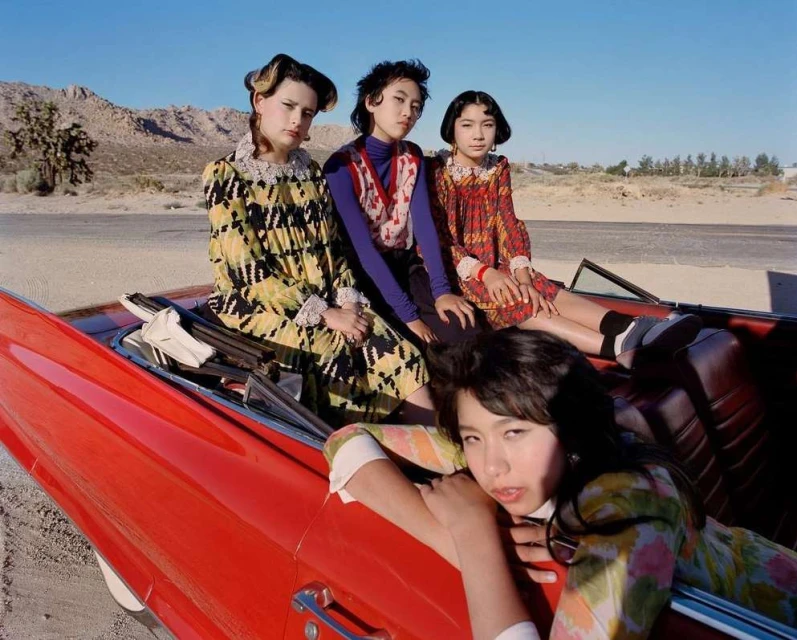 Four young women pose in a red convertible in a desert landscape, wearing colorful vintage-style dresses and hairstyles with mountains in the background.