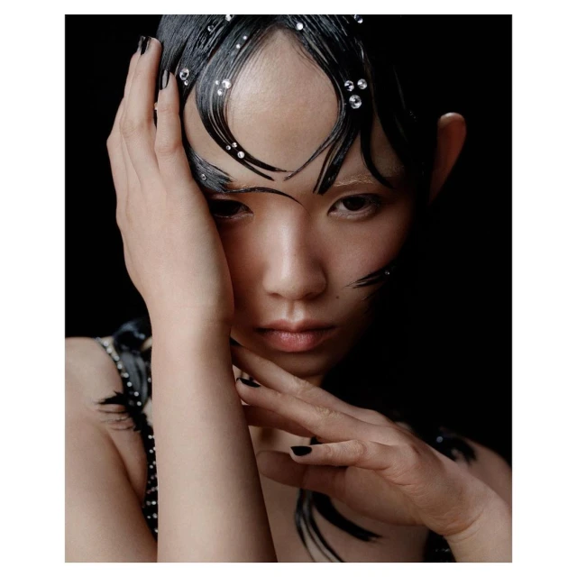 Close-up portrait of an Asian woman with black hair, embellished with shiny jewels, posing against a dark background, and wearing black nail polish.